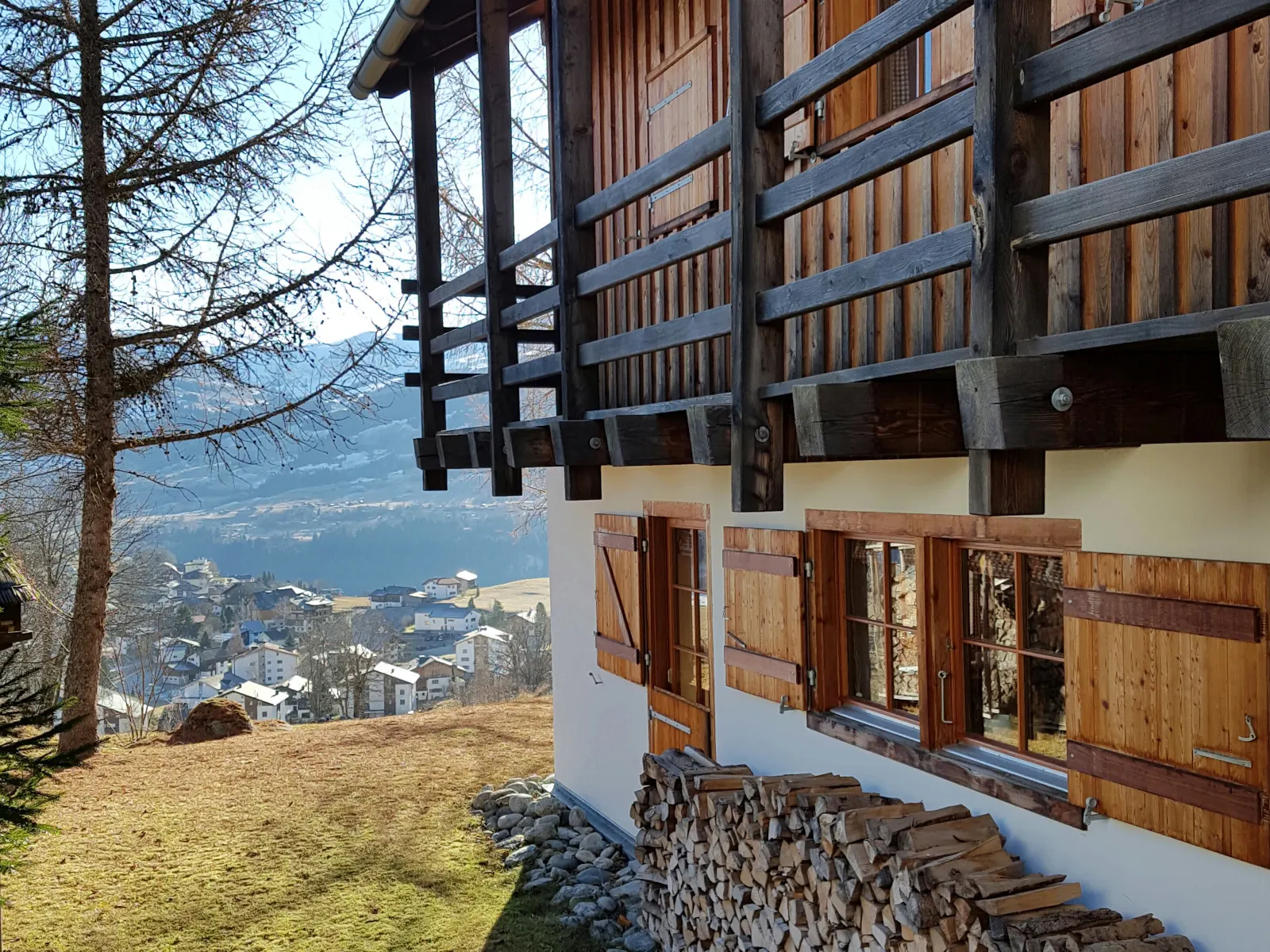 Der Balkon aus dunklem Holz, darunter gestapeltes Brennholz und der Blick hinunter ins Dorf