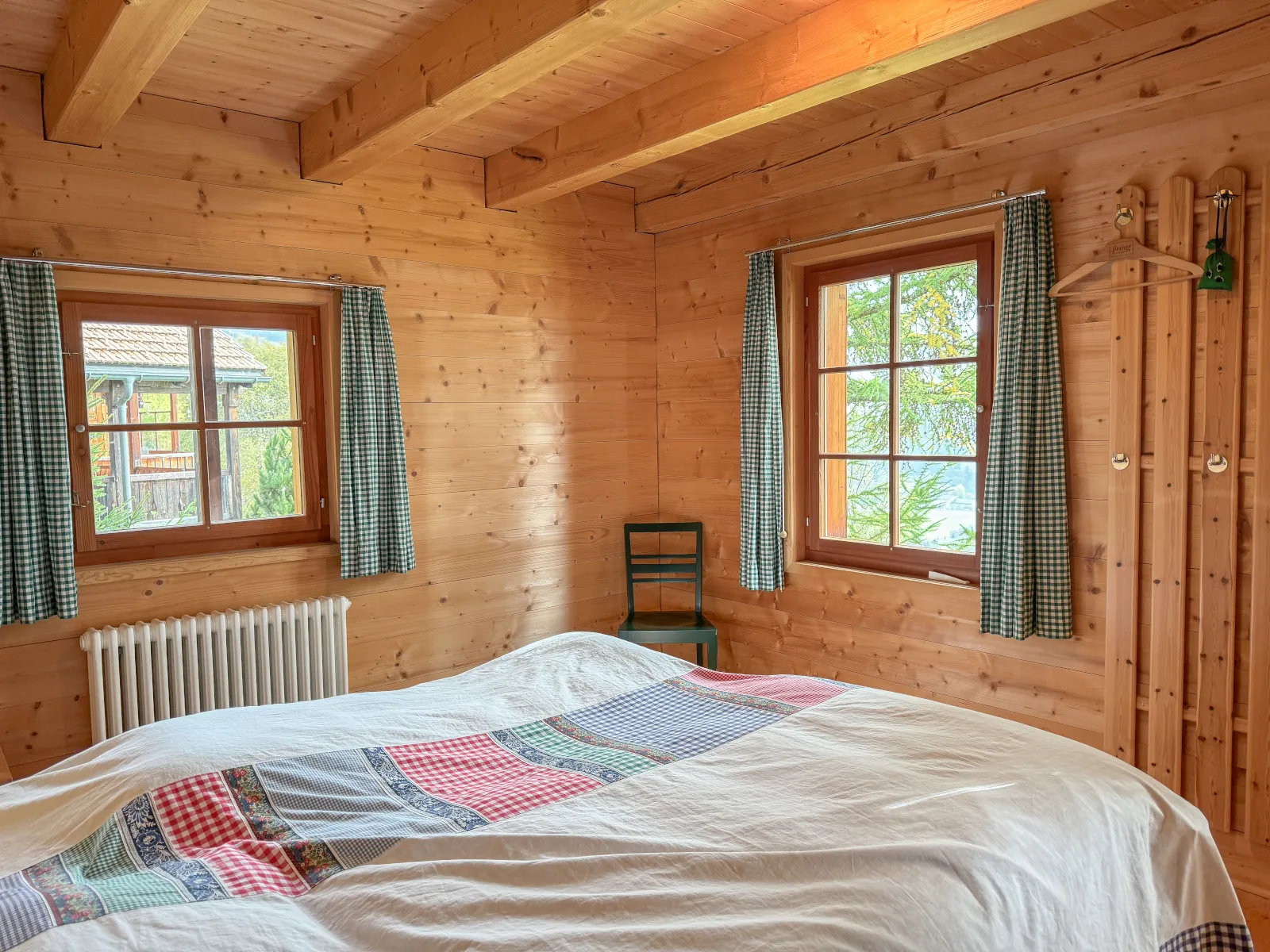 Doppelzimmer mit Patchwork-Überwurf, grün karierten Vorhängen und zwei Fenstern Richtung Lärchen